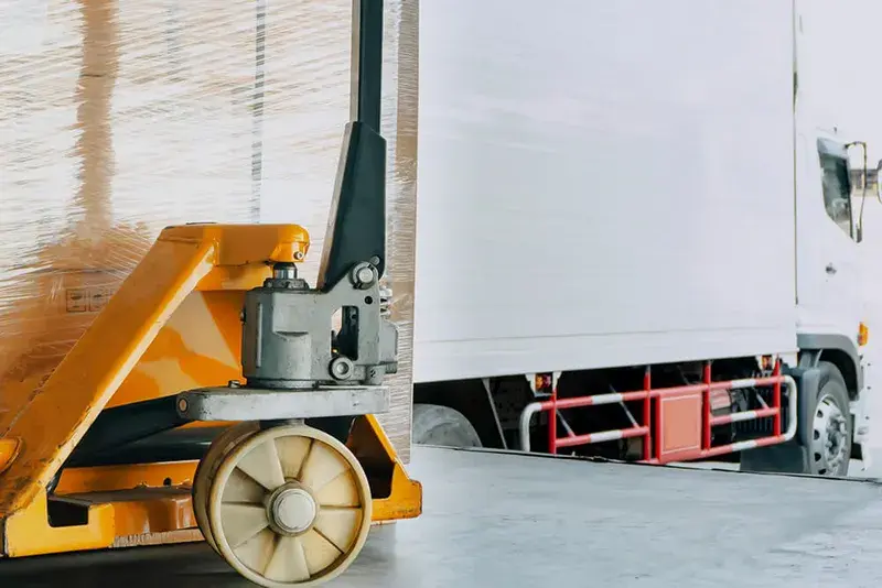Pallet jack moving shrink-wrapped freight toward a box truck at a loading dock