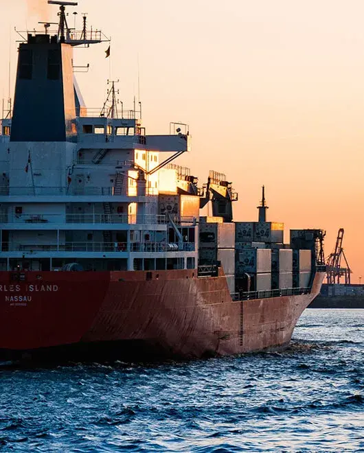 Container vessel leaving harbor at sunset with port cranes behind